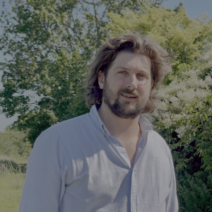 a headshot portrait of James Hare in blue shirt with a background setting of trees and blue sky